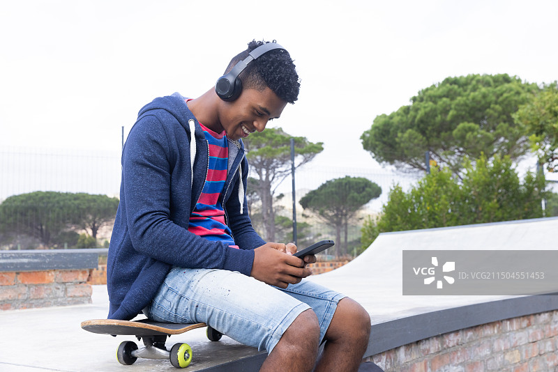 戴着耳机听音乐，坐在滑板上用智能手机的少年图片素材