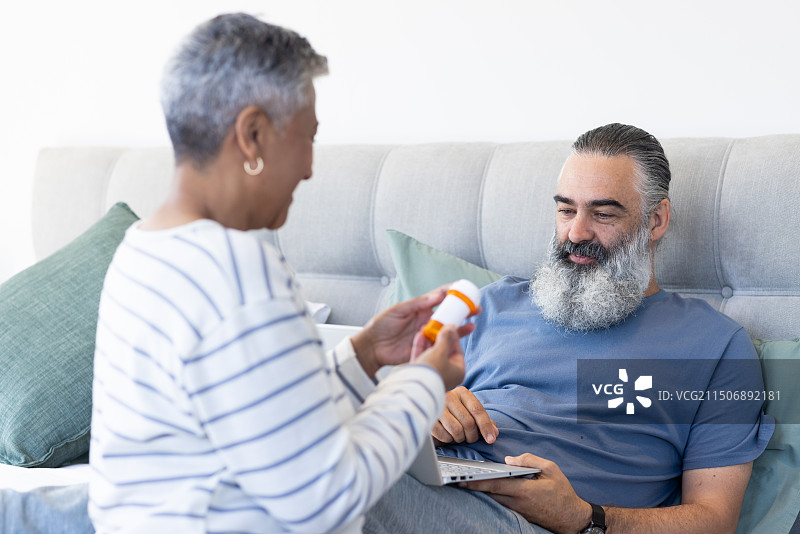 老年妇女拿着药瓶，男人在床上用笔记本电脑放松图片素材