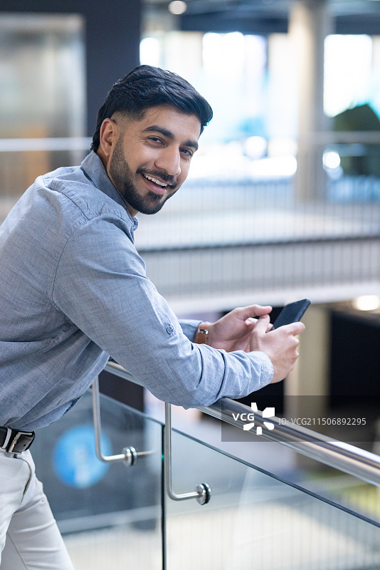 在现代办公室里倚靠栏杆手持智能手机的微笑商人图片素材
