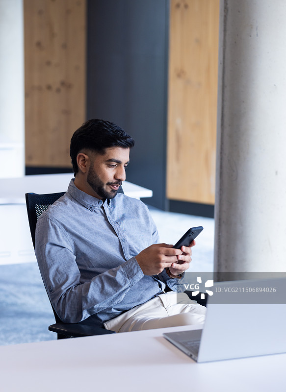 在现代办公室里，男人坐在办公桌前用手机和笔记本电脑图片素材