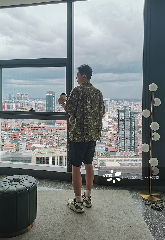 拿着咖啡在落地窗前欣赏金边城市风光的男子图片素材