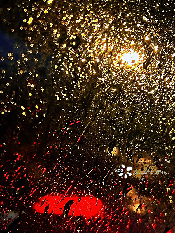 下雨的夜晚汽车玻璃窗上的雨滴映衬在车灯和路灯照射下的特写镜头图片素材