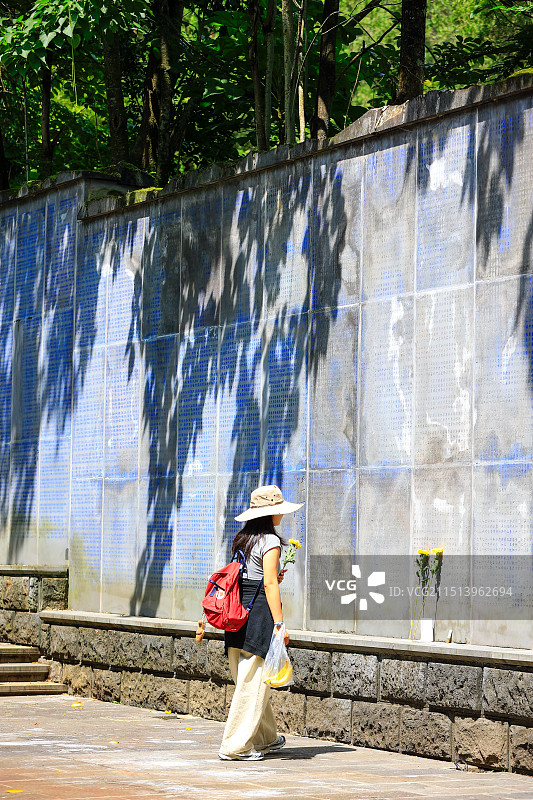 一位年轻女子站在刻满抗日战争牺牲者姓名的纪念墙前，手持鲜花表达敬意，缅怀为国捐躯的烈士图片素材