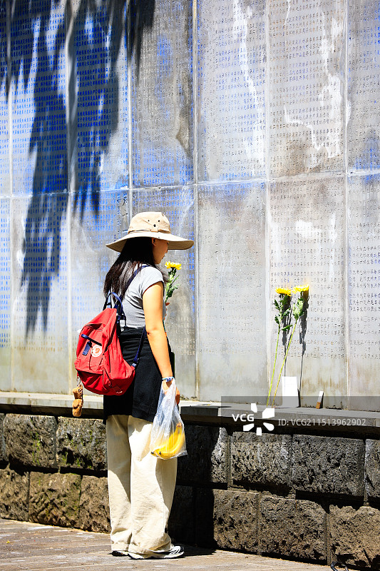 一位年轻女子站在刻满抗日战争牺牲者姓名的纪念墙前，手持鲜花表达敬意，缅怀烈士图片素材