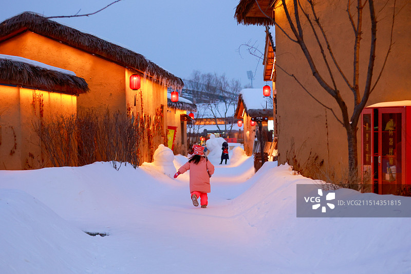 小女孩在雪地里奔跑，灯笼照亮了两旁的传统泥屋，营造出温馨的冬日氛围，充满了童趣与活力图片素材