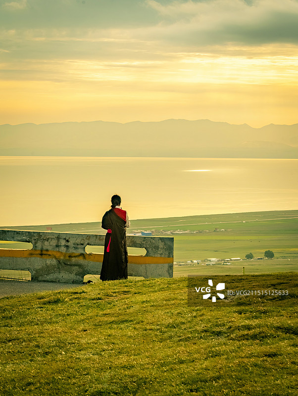 青海湖蔚蓝色湖水，嫩绿色草原，公路蜿蜒，牛羊成群，日出时分温暖的光线和彩色的朝霞，一个藏族女孩眺望图片素材