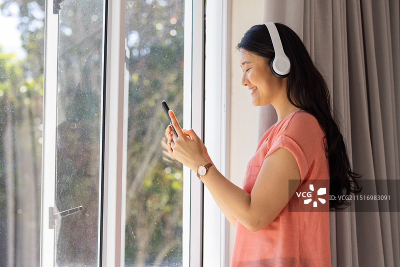 戴着耳机，站在窗边使用智能手机微笑的亚洲女人图片素材