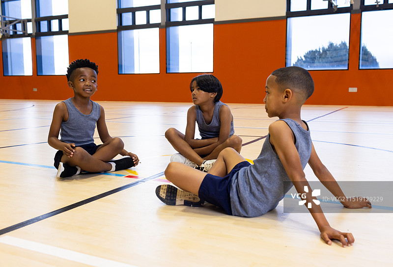 学校体育馆内，三个多种族男孩穿着运动服坐在地板上交谈图片素材