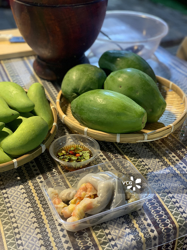 云南西双版纳星光夜市街头的当地特色小吃—卷粉，以云南特色织物、器物和青瓜青芒为背景图片素材