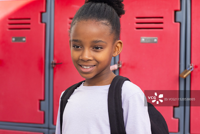 微笑的非洲裔美国女孩，背着背包站在红色学校储物柜前图片素材