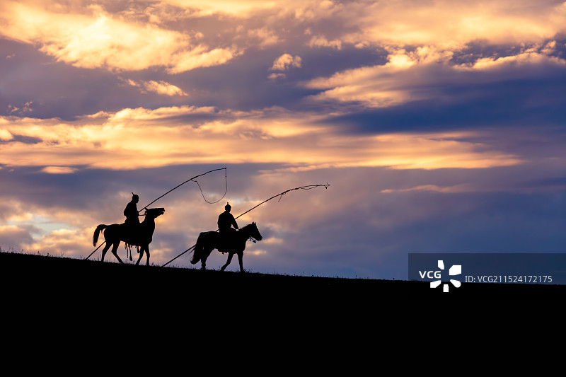 内蒙古乌兰布统草原，绚丽夕阳晚霞映衬下，天边两位身着传统蒙古族服装手持套马杆的骑马牧民的剪影图片素材