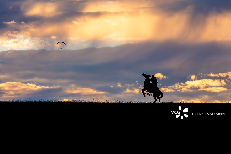 内蒙古乌兰布统草原绚丽夕阳晚霞映衬下，身着传统蒙古族服装的骑马牧民朝着天上滑翔伞，纵马而立剪影图片素材
