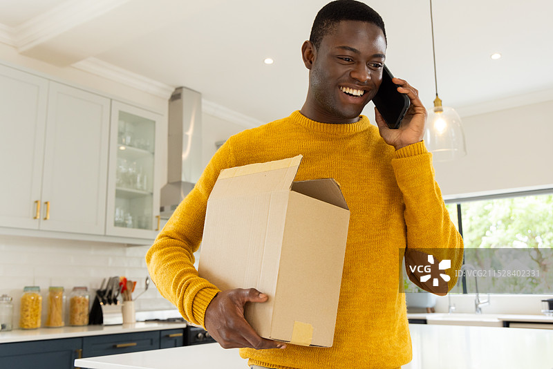 微笑的非洲裔美国男子在家中的厨房里一边打电话一边拿着纸箱。图片素材
