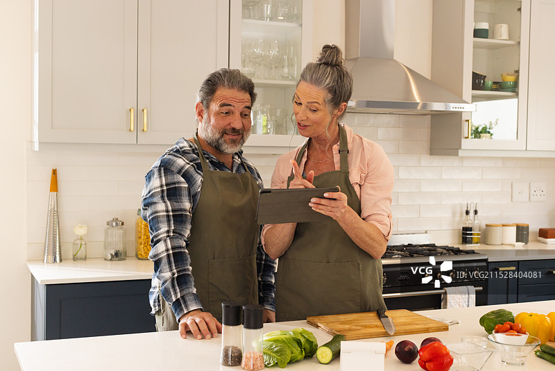 成熟夫妇在厨房使用平板电脑查看食谱，微笑并专注，居家环境。图片素材