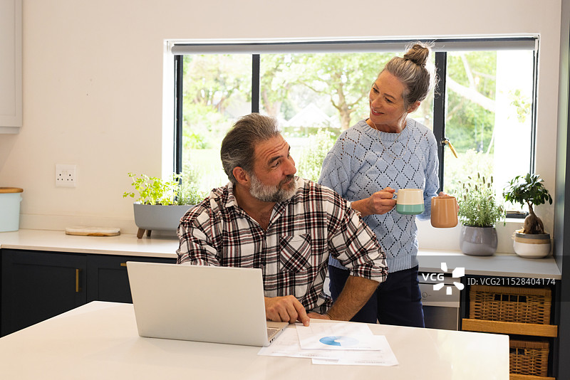 成熟夫妇在家享受咖啡时光，男人在厨房使用笔记本电脑图片素材