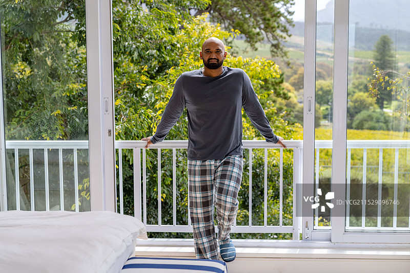 快乐的混血男性穿着睡衣，站在阳台上，背景是树梢。图片素材
