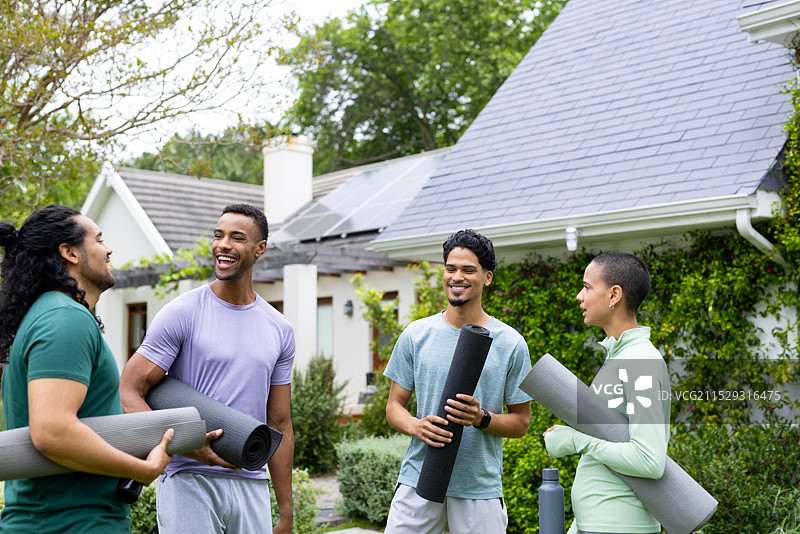 年轻的多种族朋友们在房子外面笑着，手里拿着瑜伽垫，房顶上有太阳能板。图片素材