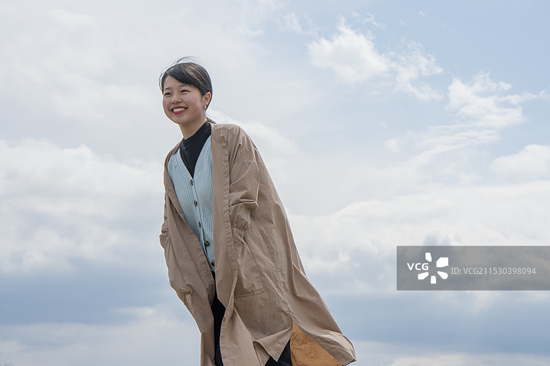 女人在广阔的天空下，笑着把手放在外套里图片素材