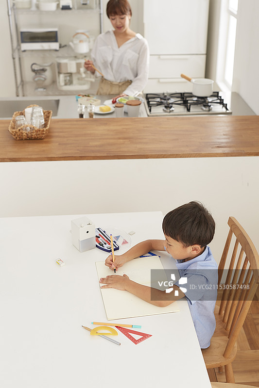 女人在厨房做午餐，男孩在餐厅学习。图片素材