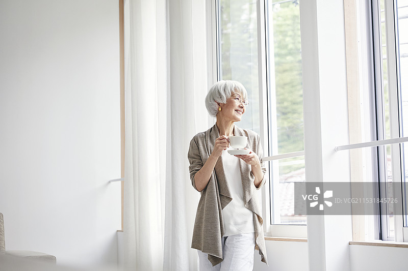 一位老年女性站在窗边，手中拿着杯子图片素材
