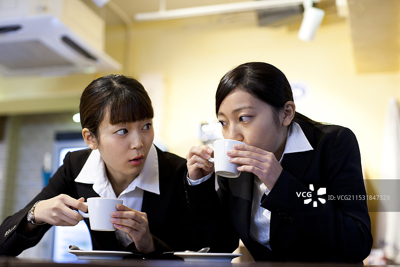 两位穿着西装的女性在咖啡馆的柜台上喝着加了大量奶泡的咖啡。图片素材