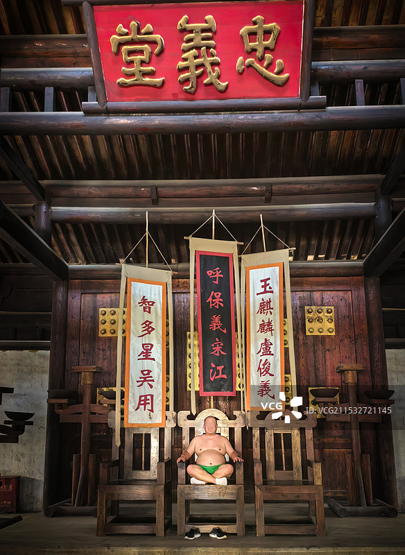 梁山忠义堂大堂内，正面三把金交椅挂着宋江吴用卢俊成的旗幅，中间椅子上一个弥勒佛一样的游客端坐如佛像图片素材