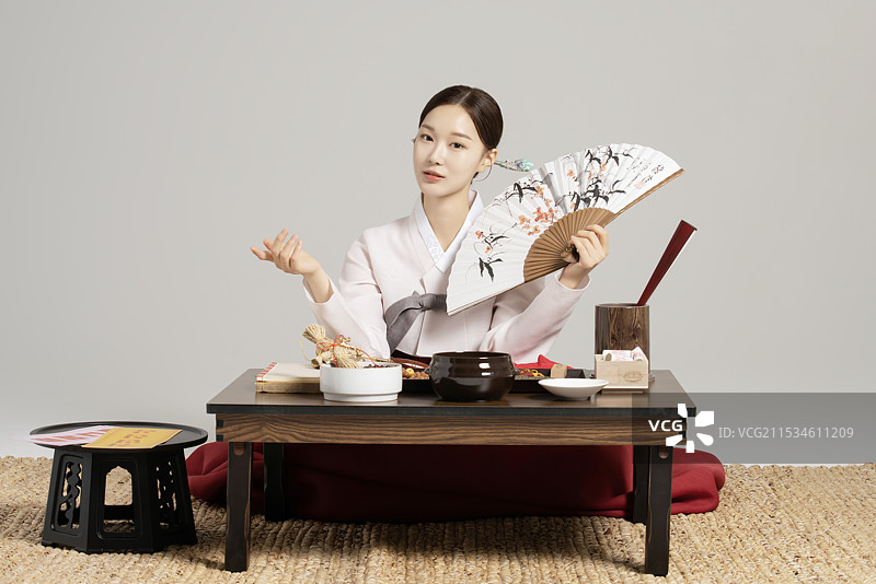 一位年轻的萨满穿着韩服，手持扇子，站在一张摆满萨满工具的桌子前。图片素材
