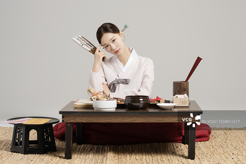 一位年轻的萨满穿着韩服，手持扇子，思考着面前摆满萨满工具的桌子。图片素材