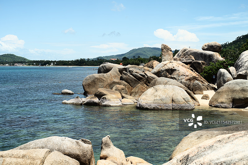 泰国苏梅岛海水清澈，岸边巨石嶙峋，远处山峦起伏，蓝天白云映衬，展现出一幅宁静而壮美的海滨风光。图片素材
