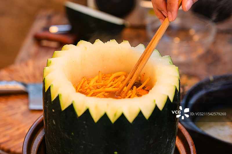 一个被挖空的绿色冬瓜盅，内部装着热气腾腾的食物，南瓜边缘呈锯齿状，背景有厨具，透着创意烹饪的氛围。图片素材