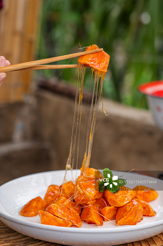 一只筷子夹着一块橙红色的拔丝地瓜，食物上有糖浆拉丝，放在一个白色盘子里，盘子上还点缀着一片绿叶。图片素材