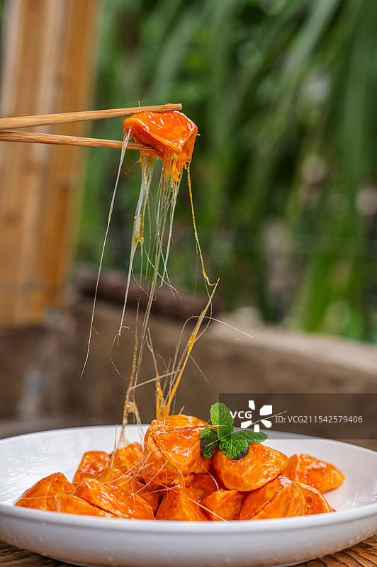 一只筷子夹着一块橙红色的拔丝地瓜，食物上有糖浆拉丝，放在一个白色盘子里，盘子上还点缀着一片绿叶。图片素材
