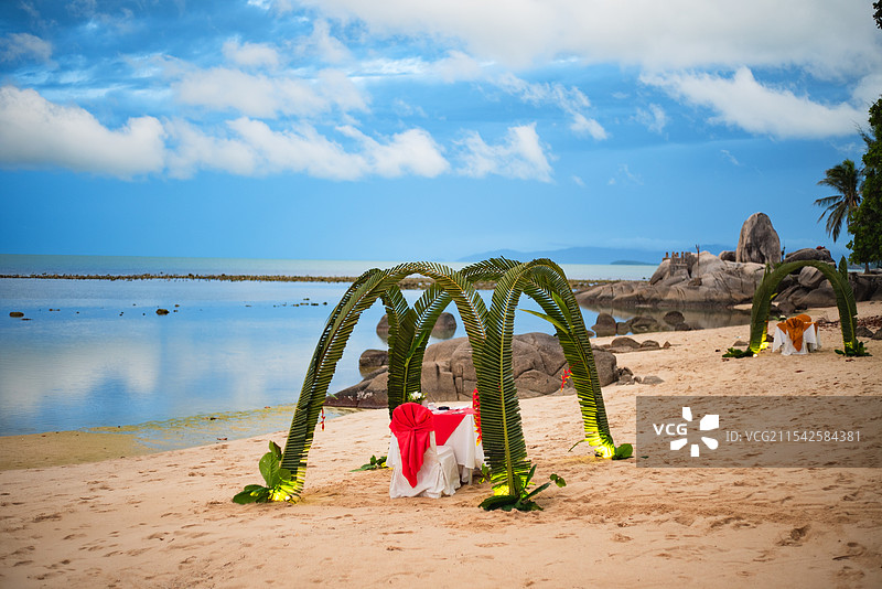 沙滩上有一个由绿植搭建的拱门，拱门下摆放着红白色桌椅，背景是平静的海面和蓝天白云，充满浪漫氛围。图片素材