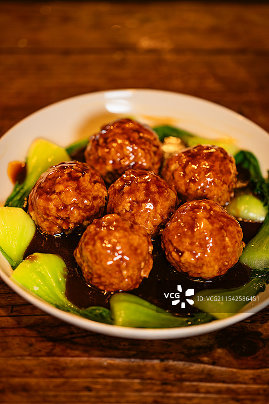 一个白色盘子里装着几个色泽诱人的肉丸，肉丸周围点缀着绿色的青菜，看起来很美味。图片素材