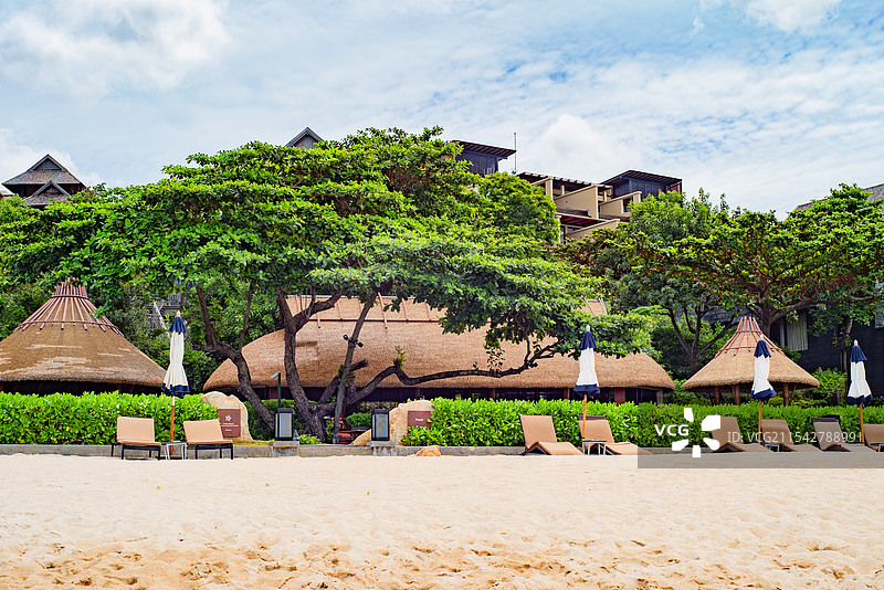 一片沙滩，沙滩上整齐排列着躺椅和遮阳伞，周围绿树环绕，蓝天白云，尽显休闲度假氛围。图片素材