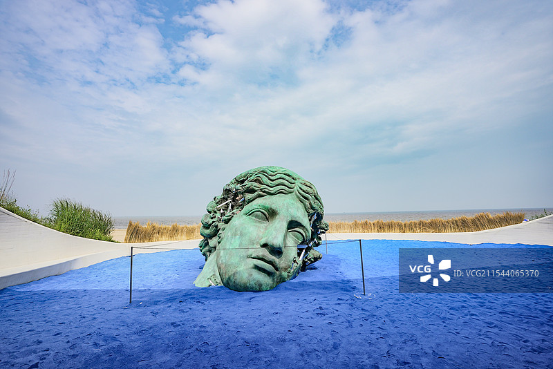 一个巨大的青铜色雕塑头像，置于一片蓝色的沙地上，背景是蓝天白云和大海，雕塑周围有围栏，整体场景图片素材