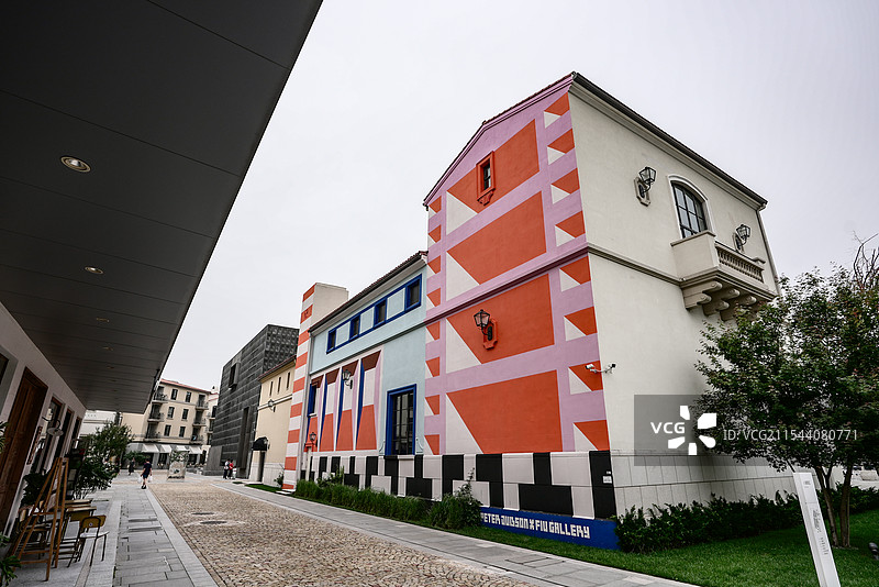 栋建筑，外墙色彩丰富，有红、白、粉、蓝等颜色，搭配几何图案，周围有绿植、石板路，远处还有其他建筑，颇图片素材