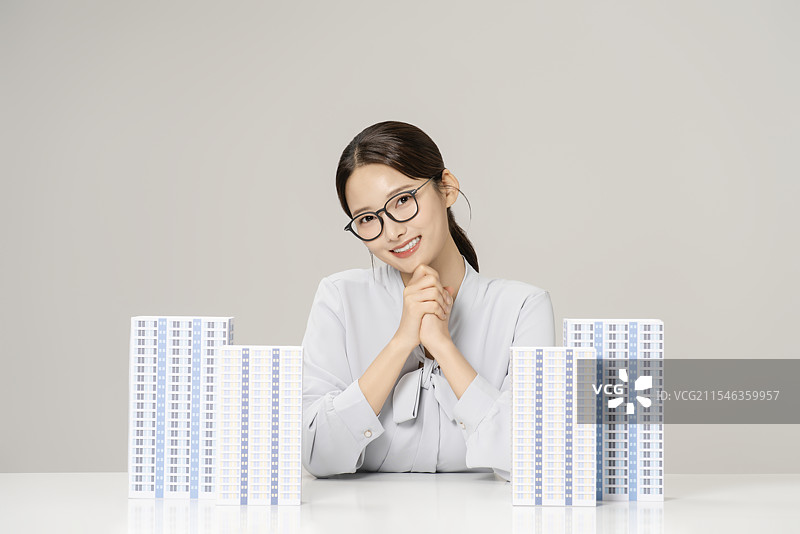一位20多岁的年轻女性坐在桌子前，桌上摆放着一个公寓模型。图片素材