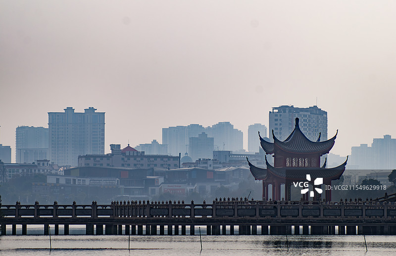 吉安庐陵文化生态园，中式古亭立于水上长桥，远处现代高楼在朦胧雾气中若隐若现。图片素材
