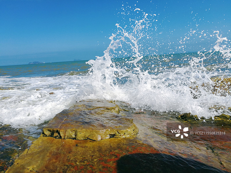 蓝色天空背景下，海浪冲击岩石，浪花翻滚图片素材