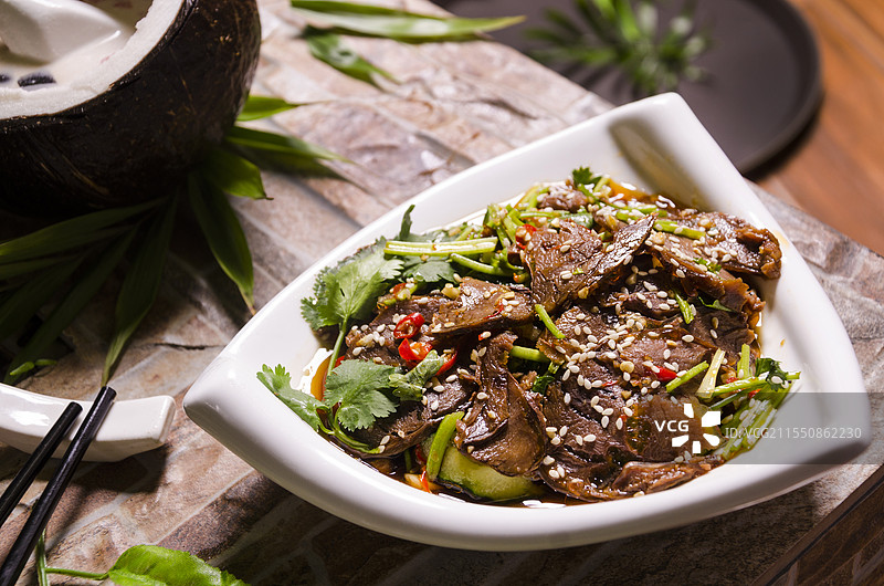 一盘撒有香菜、芝麻、辣椒等佐料的凉拌牛肉，背景有切开的椰子和绿叶。图片素材