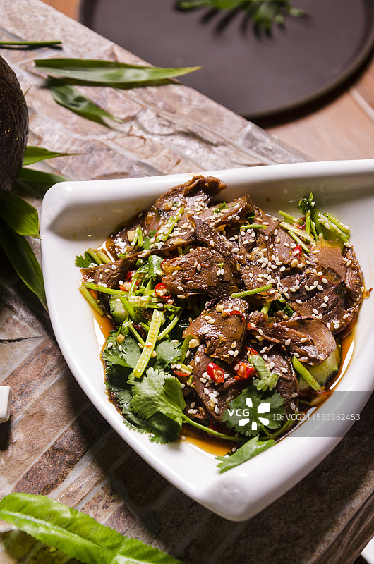 一盘撒有香菜、芝麻、辣椒等佐料的凉拌牛肉，背景有切开的椰子和绿叶。图片素材