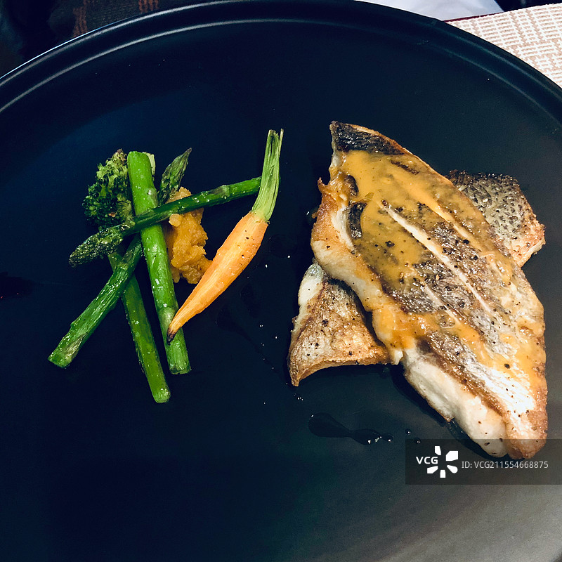 精致的香煎海鱼搭配清新的芦笋和脆嫩的胡萝卜放在黑色的餐盘上，淋上浓郁酱汁，色香味俱佳。图片素材