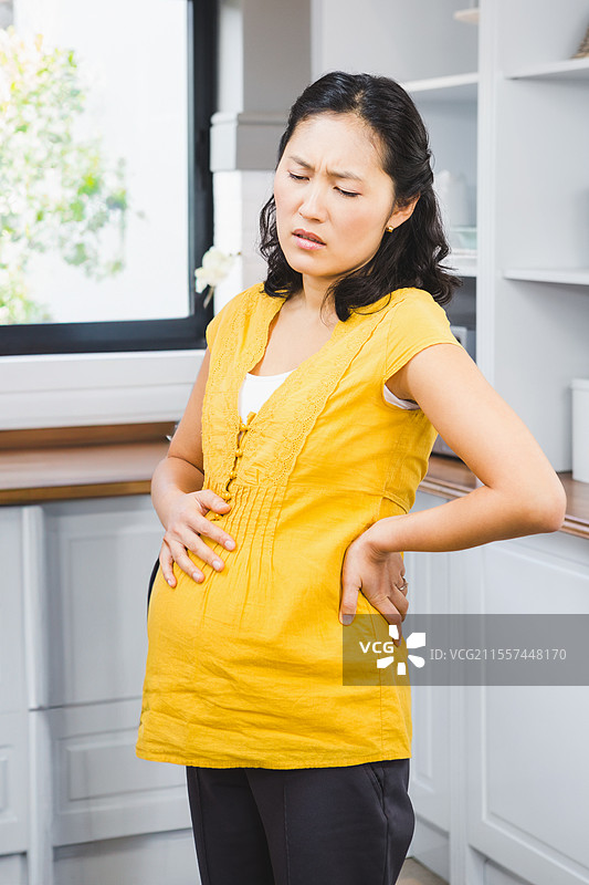 怀孕的亚洲女性在厨房感到不适，双手抱住腹部。图片素材
