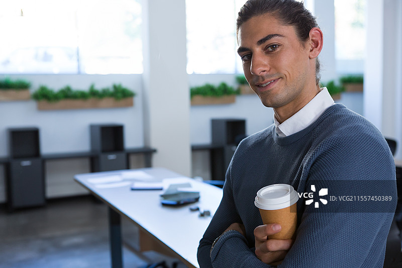 自信的男人手持咖啡杯，站在现代办公室里，温暖地微笑，留有空白空间图片素材