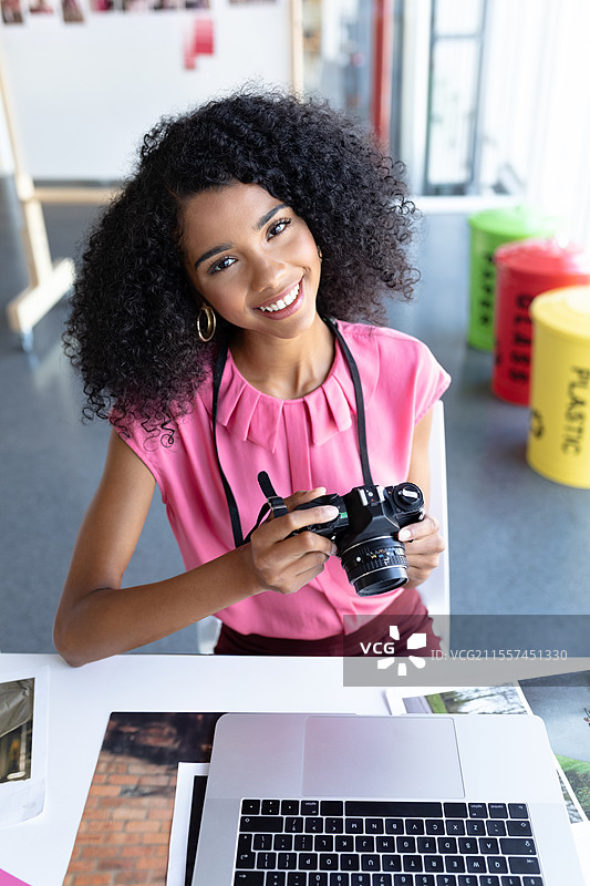 微笑的女性在办公桌前拿着相机，桌上有笔记本电脑和照片。图片素材
