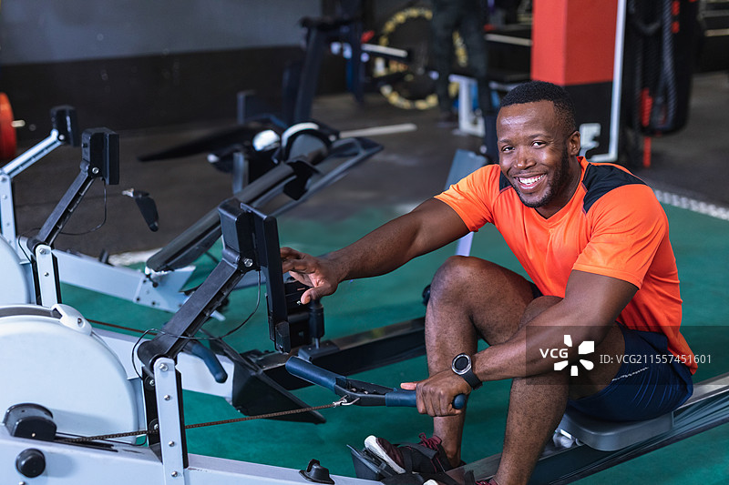 穿着橙色衬衫的男子在健身房使用划船机，面带微笑，享受锻炼。图片素材