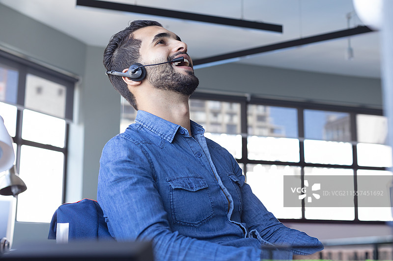 在办公室桌前，戴着耳机的商人一边工作一边笑着。图片素材