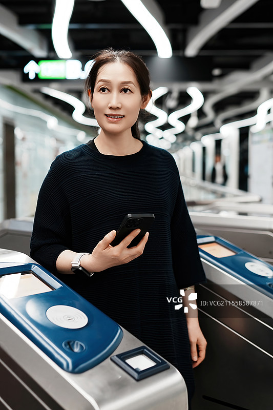 在现代城市环境中，年轻女子手持智能手机站在地铁旋转栅门前图片素材