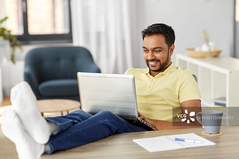 科技、远程工作和生活方式概念 - 开心微笑的印度男子在家办公时将脚放在桌子上，使用笔记本电脑。男子在家中用笔记本电脑休息，脚放在桌子上。图片素材